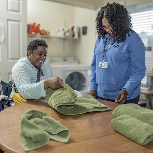 CNS patient folding laundry in residential rehab while NRS staff looks on.